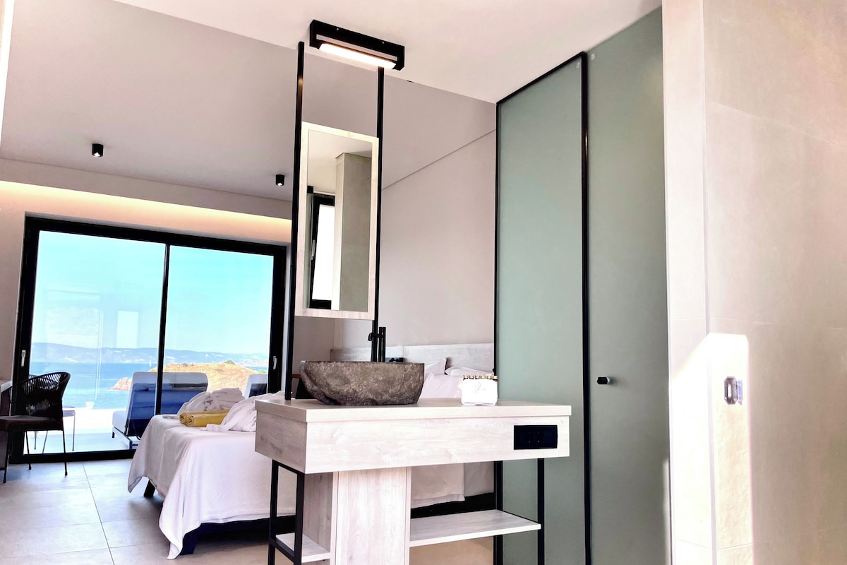 A modern hotel room with a large bed, a bedside table with a mirror, a bathroom with a large mirror, and a view of the sea.