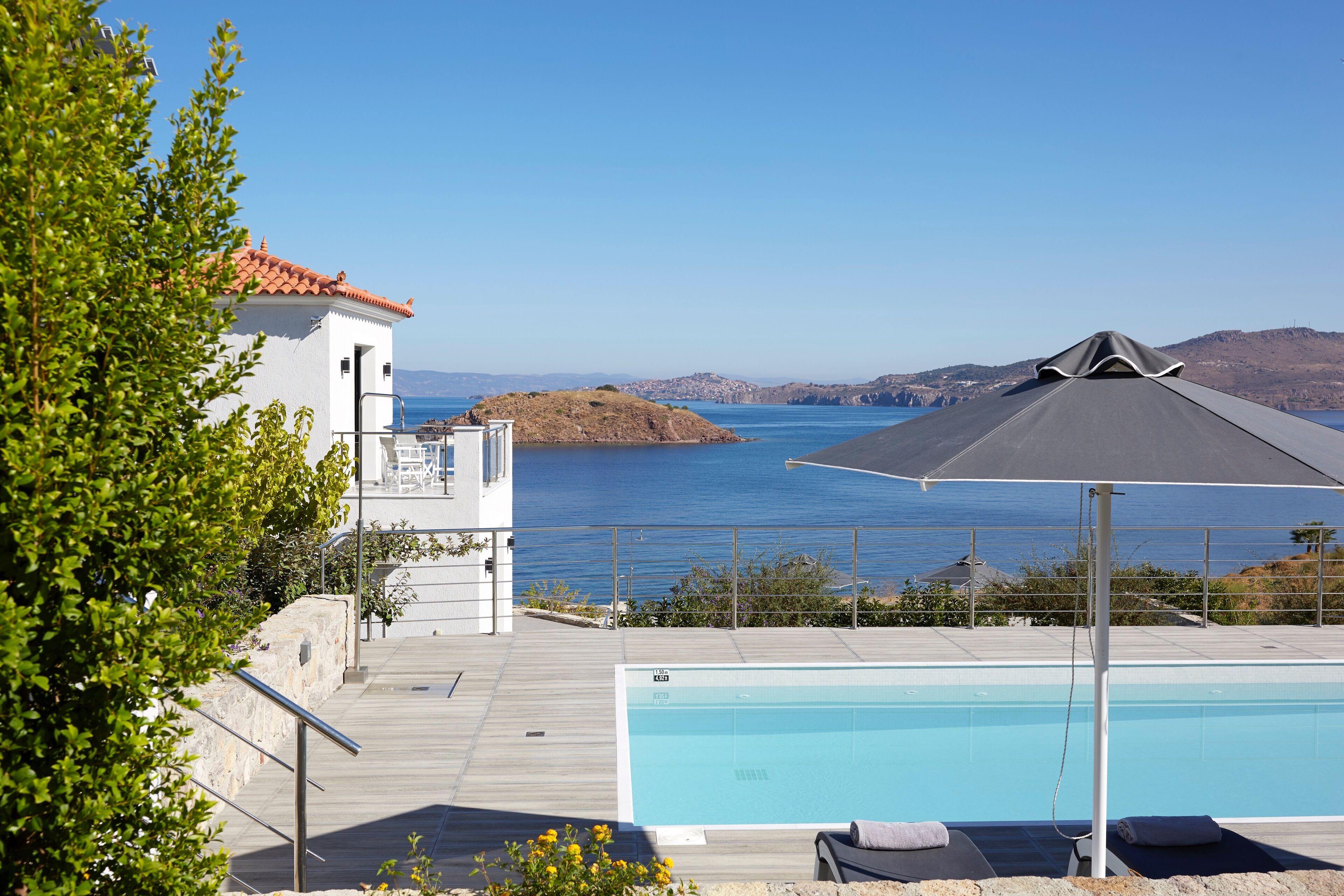 A pool area with a view of the sea, a white building with a red roof, and a wooden deck.