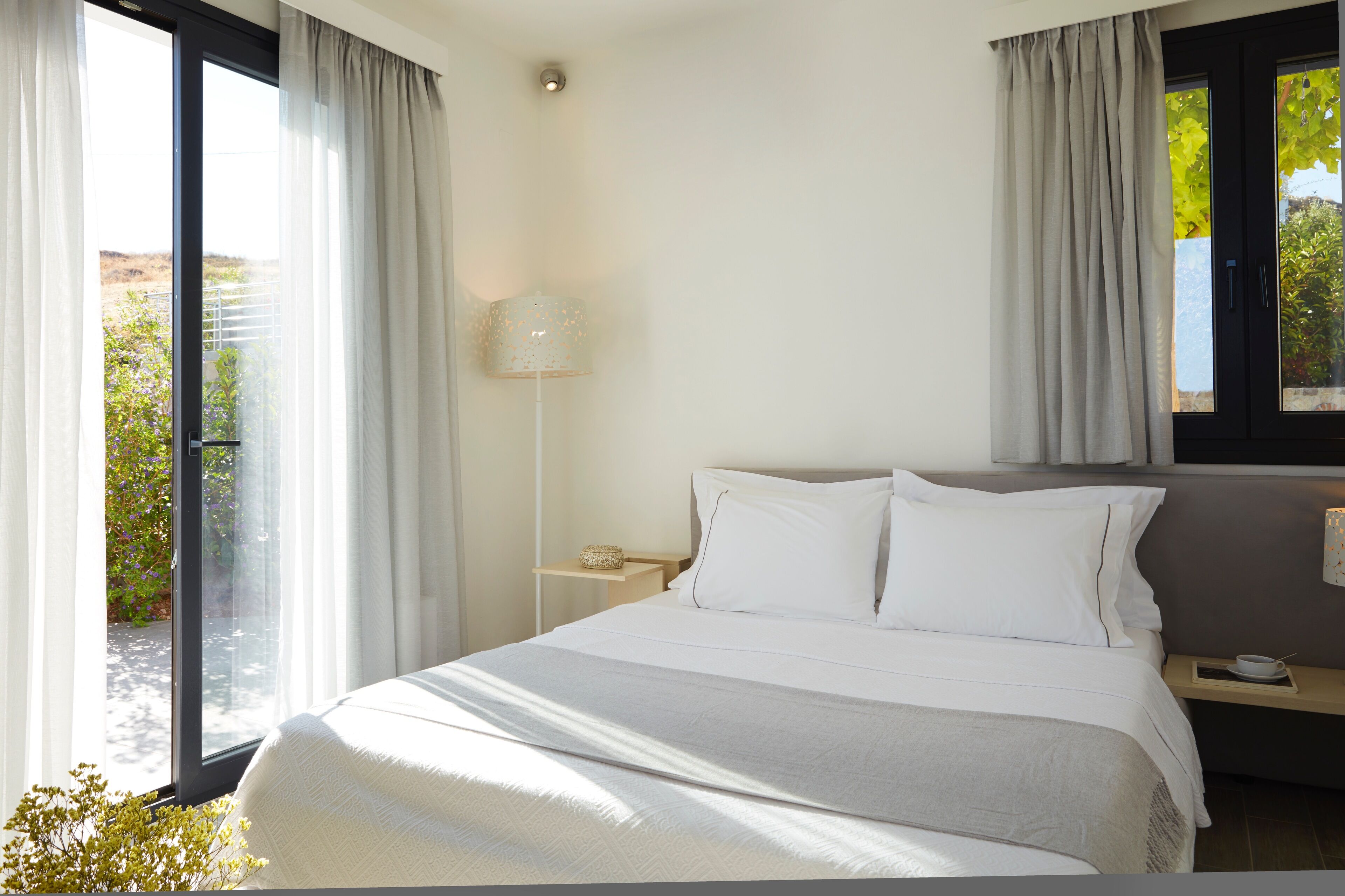 A bedroom with a large bed, a nightstand, a lamp, and a view of the outdoors.