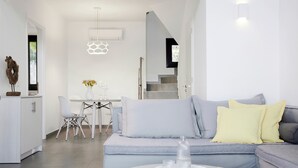 A modern living room with a grey sofa, a round glass coffee table, and a dining area with a white table and chairs. - Eleia Seafront Rooms & Villas (Lesvos)