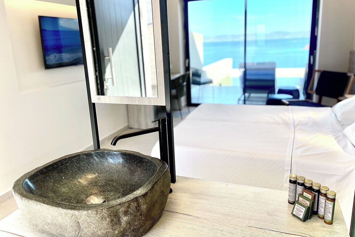 A modern hotel room with a stone basin, a bed, and a view of the sea through large windows.