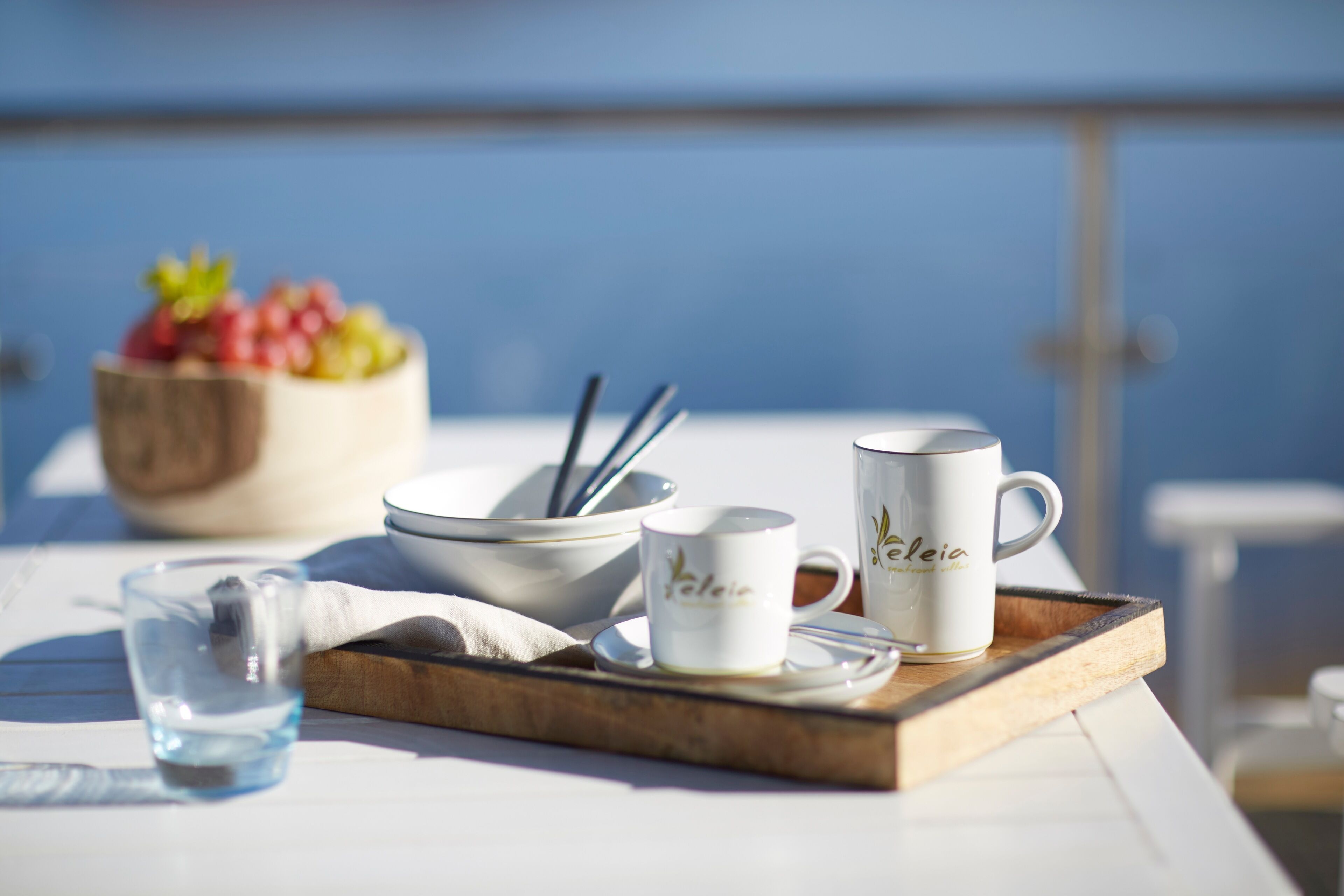 A table with a tray containing a glass of water, a white mug with "Yeleia" written on it, and a bowl with fruits and utensils.