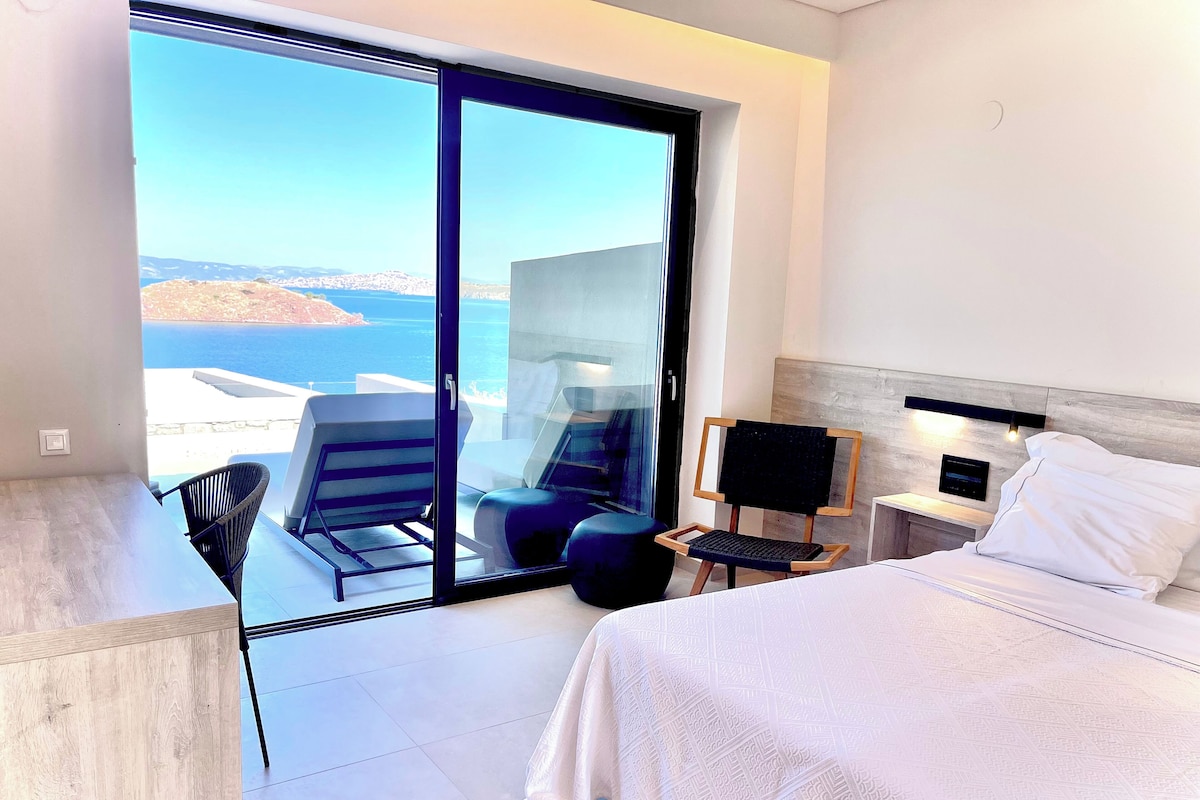 A hotel room with a bed, a desk, a chair, and a view of the sea.