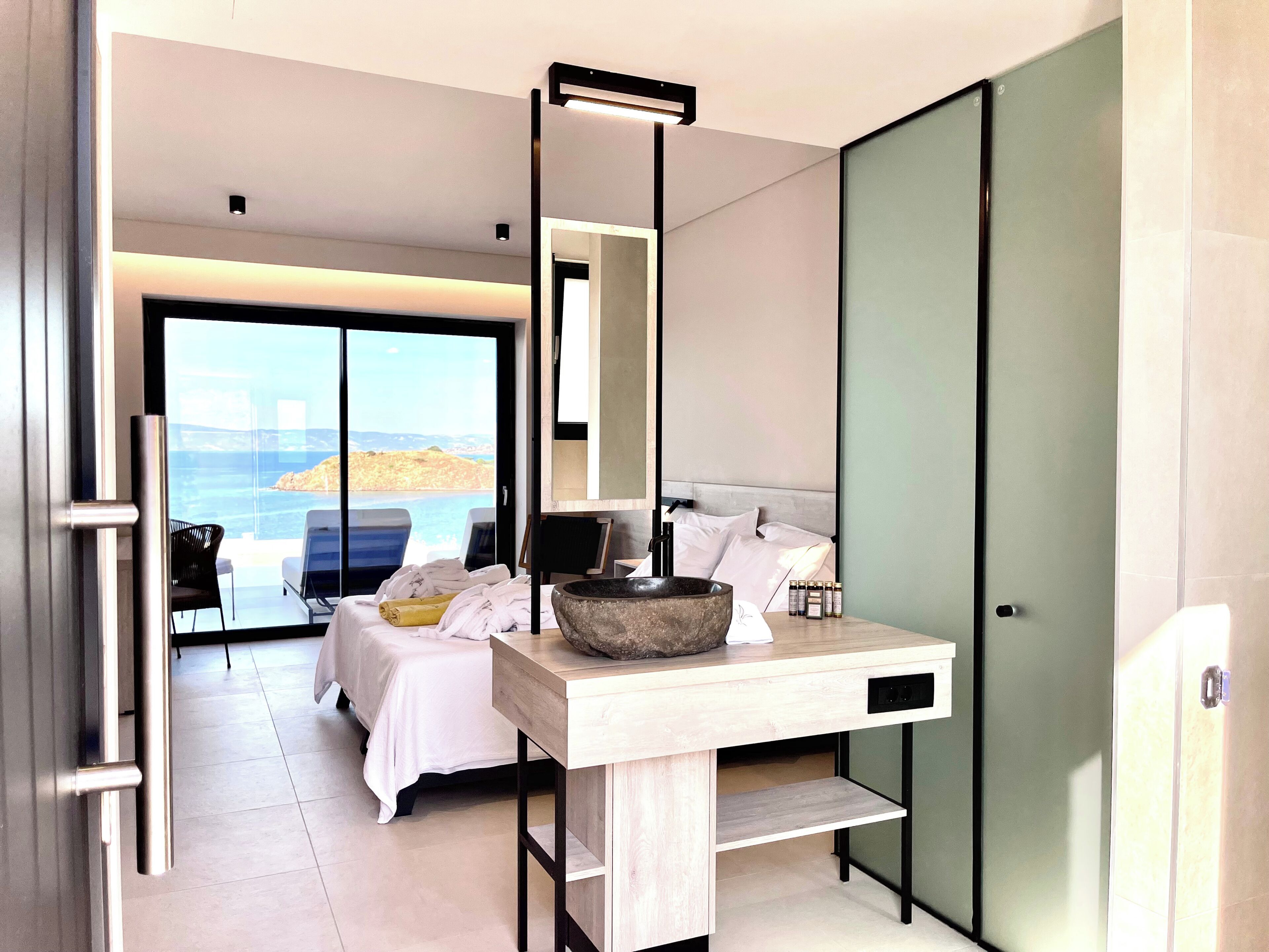 A modern hotel room with a bed, a wooden desk, a large stone bowl, and a view of the sea.