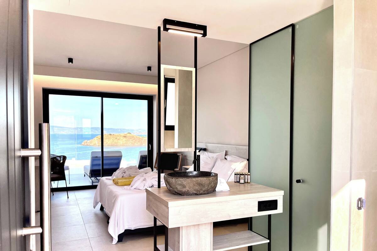 A modern hotel room with a bed, a wooden desk, a large stone bowl, and a view of the sea.