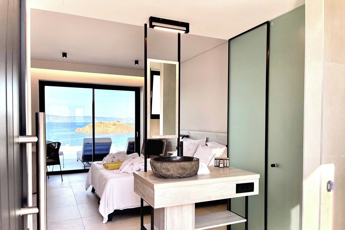 A modern hotel room with a bed, a wooden desk, a large stone bowl, and a view of the sea.