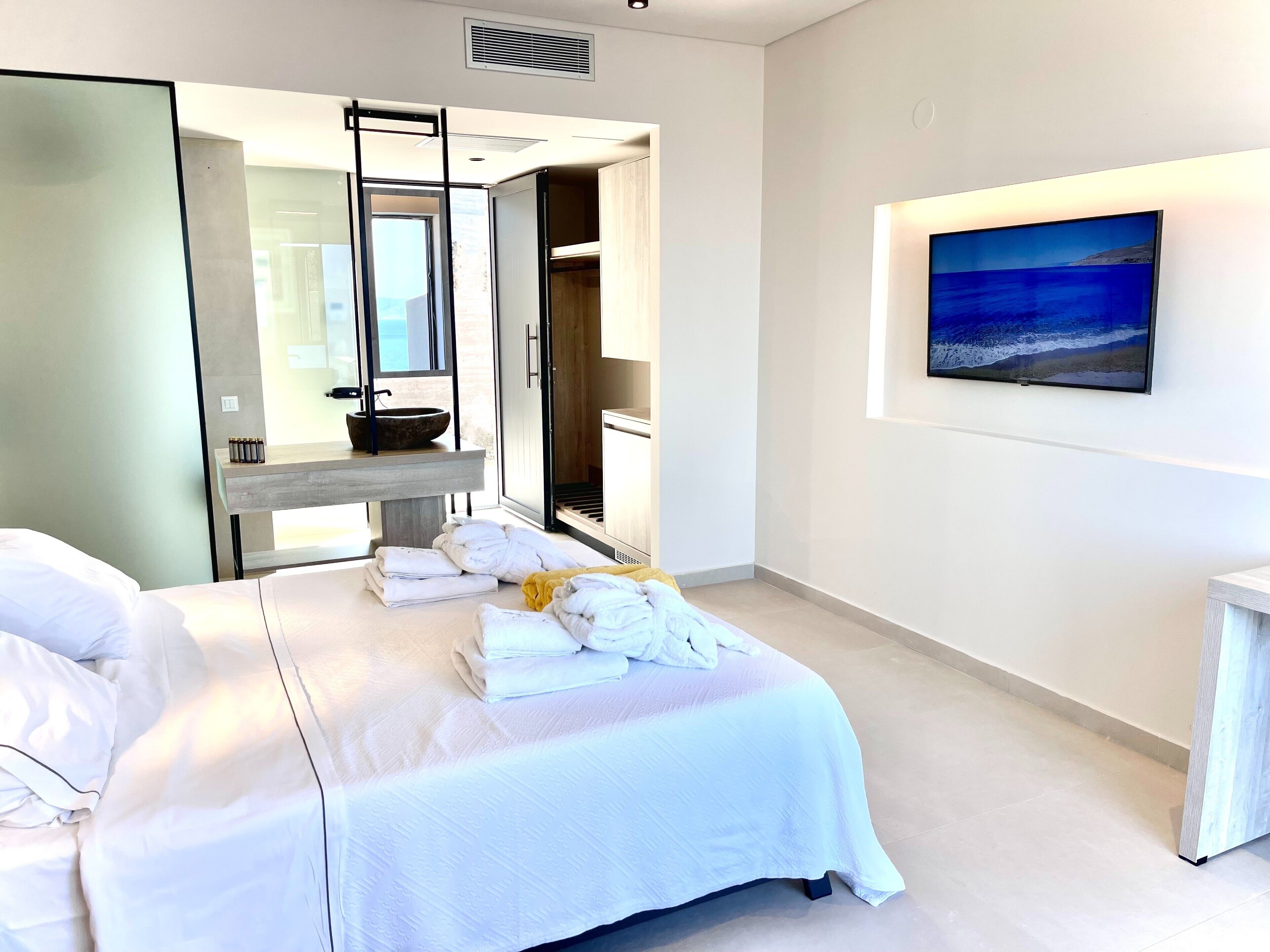 A modern hotel room with a bed, a large window, a bathroom with a sink and mirror, and a painting on the wall.