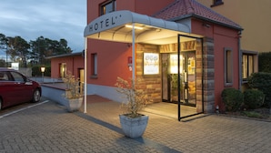 A hotel entrance with a covered walkway, a red car parked outside, and a sign indicating the hotel's name. - Le Verger De Bischwiller (Bischwiller)