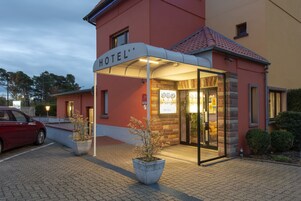 Entrada de um hotel com uma cobertura, um carro vermelho estacionado na frente e uma placa com o nome do hotel.
