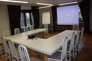Un salón de conferencias con una larga mesa, sillas blancas, una pantalla de proyector y una pizarra.