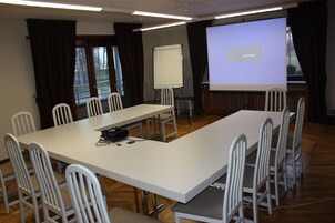 Uma sala de conferências com uma mesa comprida, cadeiras brancas, uma tela de projetor e um quadro branco.