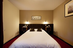 Quarto de hotel com uma cama grande, duas mesas de cabeceira com luminárias, um quadro na parede e um tapete vermelho.