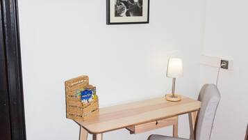 A small room with a wooden desk, a grey chair, a brown ottoman, a television mounted on the wall, and a framed picture above the desk.