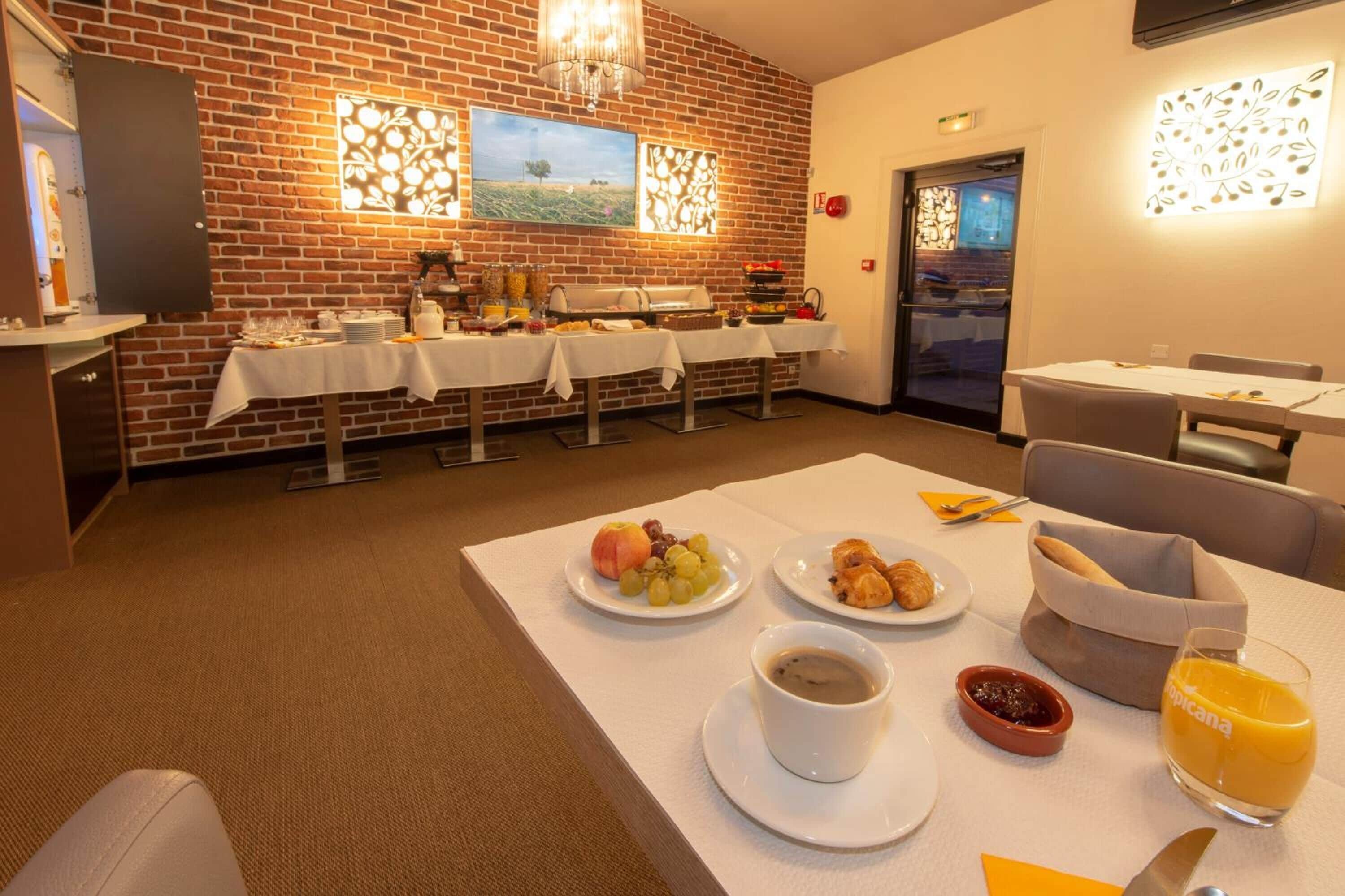 Une table dressée pour le petit-déjeuner, avec des assiettes de fruits, du café et du jus, et un buffet en arrière-plan.