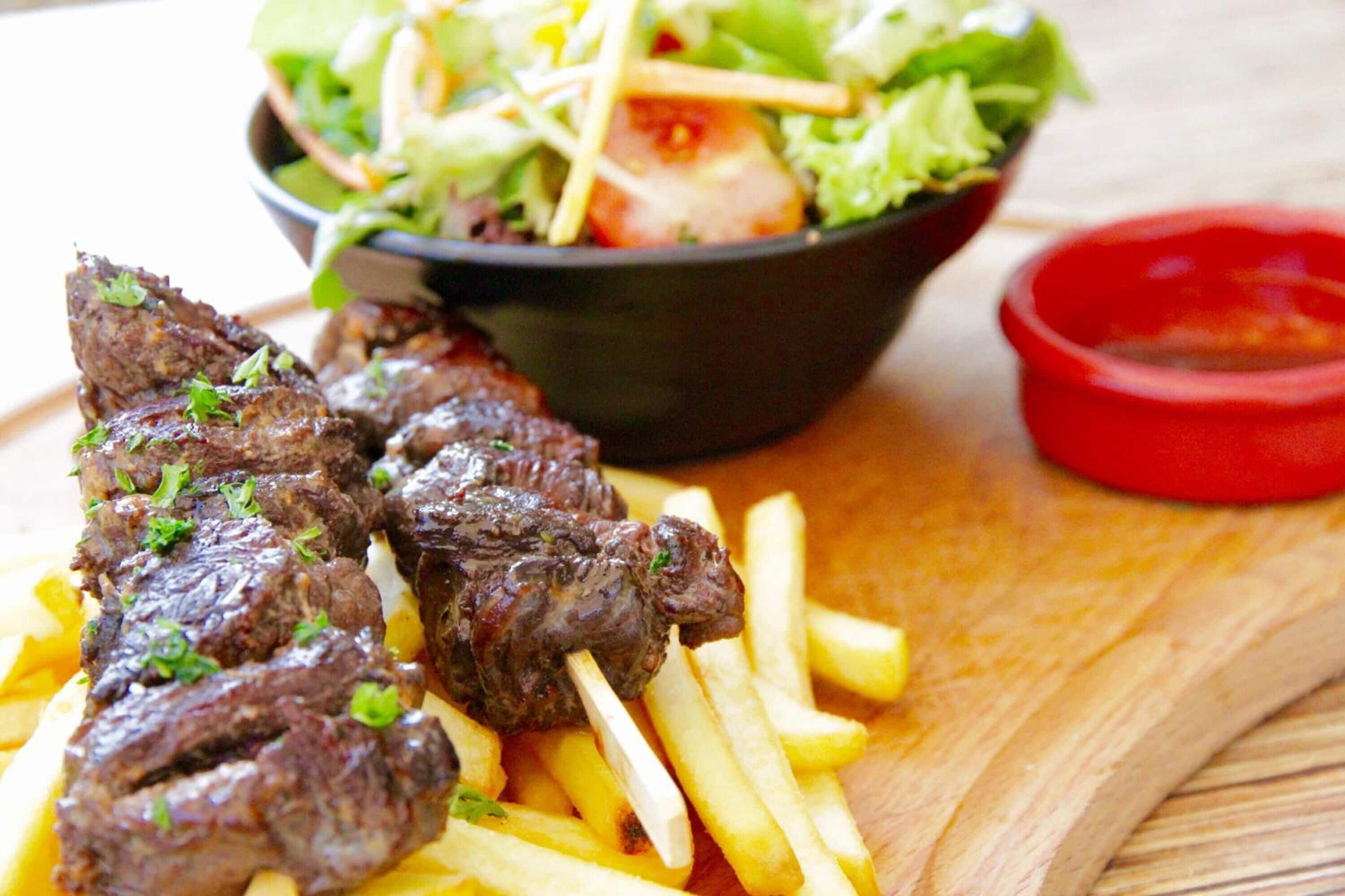 Um prato com espetinhos de carne grelhada e batatas fritas, uma salada e um pequeno recipiente vermelho com molho.