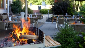 An outdoor seating area with tables and chairs, a fire pit with burning wood, and a building in the background. - Le Verger De Bischwiller (Bischwiller)