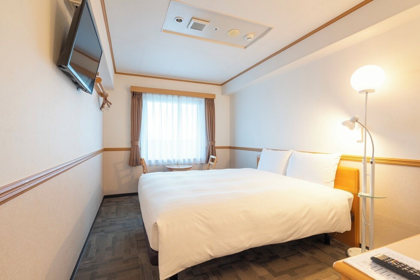Una habitación de hotel con cama, escritorio con lámpara, una silla, ventana con cortinas y televisión montada en la pared.