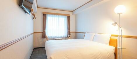 Una habitación de hotel con cama, escritorio con lámpara, una silla, ventana con cortinas y televisión montada en la pared.