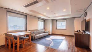A modern hotel room with a dining table, sofa, and large windows offering city views.