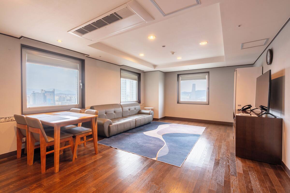 A modern hotel room with a dining table, sofa, and large windows offering city views.