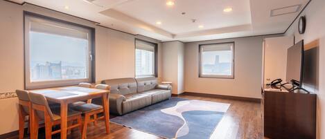 A modern hotel room with a dining table, sofa, and large windows offering city views.