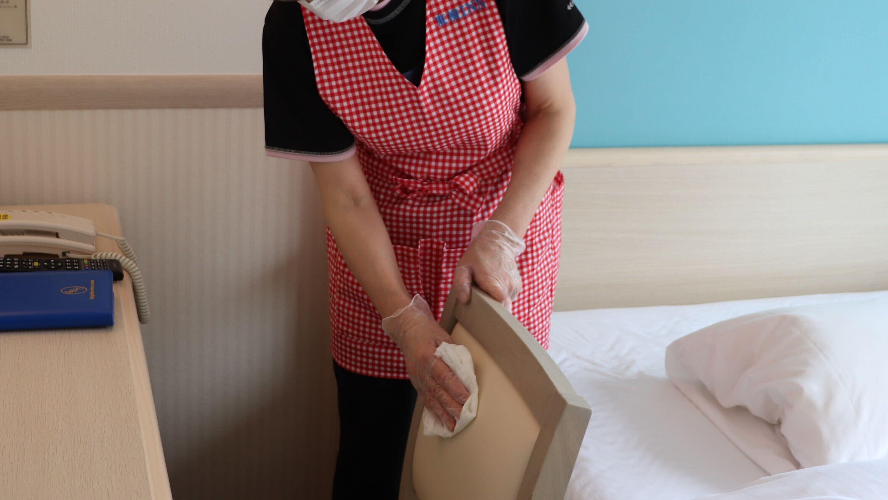 En person med ett rött och vitt rutigt förkläde som städar en säng i ett hotellrum.