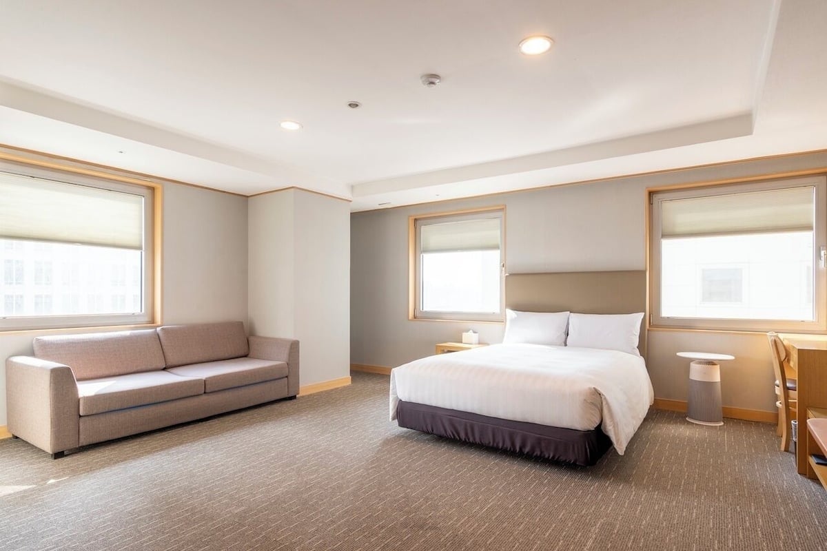 A hotel room with a bed, a sofa, a bedside table, a lamp, and large windows.