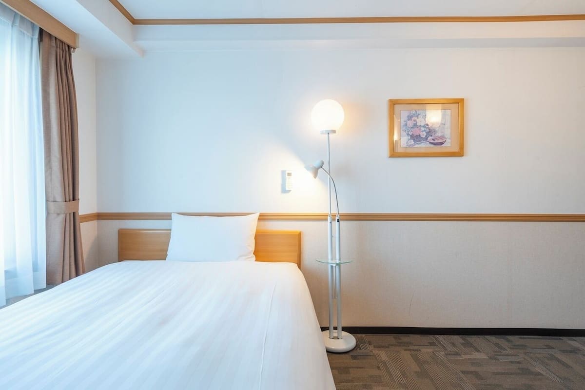 A hotel room with a bed, a bedside lamp, a framed picture on the wall, and a window with curtains.