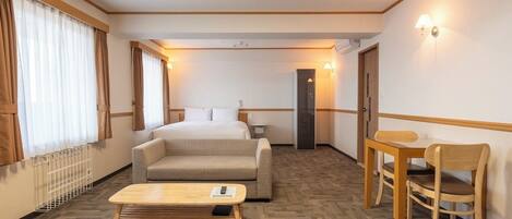 A hotel room with a bed, a sofa, a small table, and a chair. There is a radiator on the wall and a door in the background.
