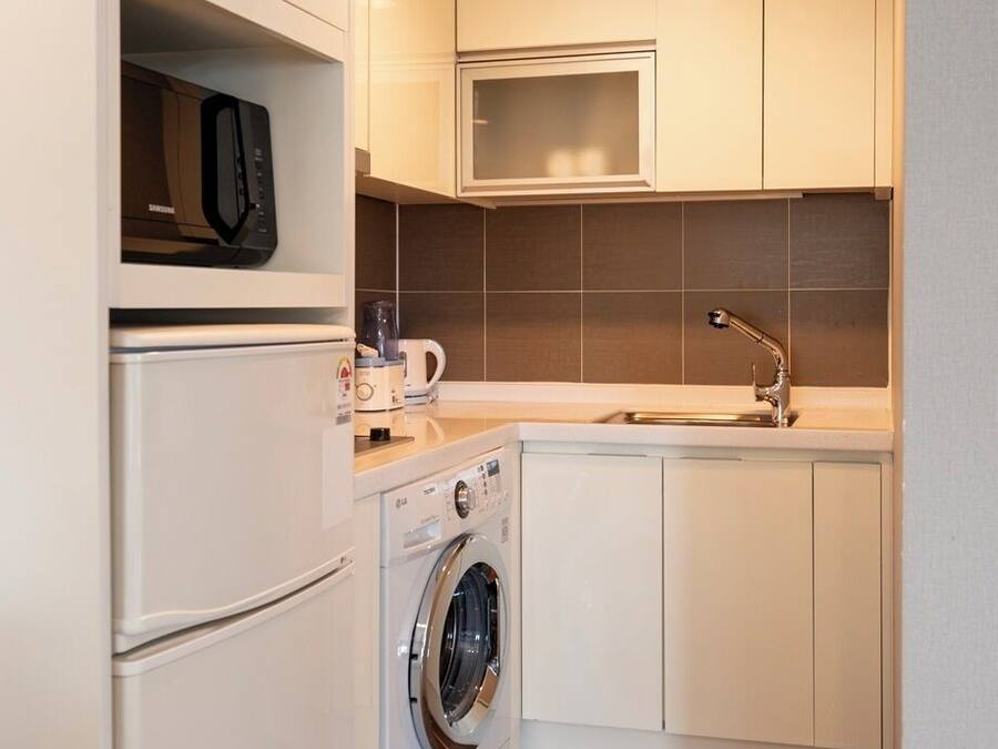 A compact kitchen with a microwave, washing machine, sink, and cabinets.