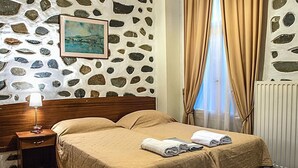 Habitación de hotel con pared de diseño de piedra, cama con colcha color beige, mesita de noche con lámpara y ventana con cortinas color beige.