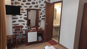 Habitación compacta con pared de diseño de piedra, un escritorio de madera con una silla, un televisor montado sobre el escritorio, un refrigerador pequeño y un baño visible a través de una puerta abierta.