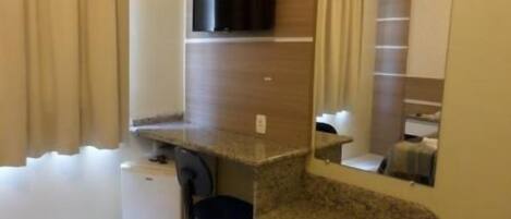 A compact room with a kitchenette, a small desk, a chair, and a mirror.
