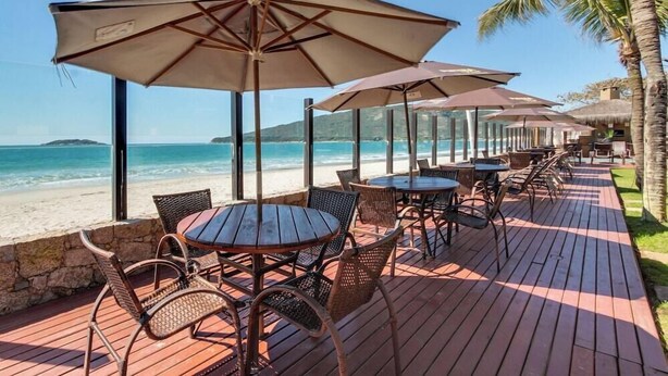 A deck with tables and chairs, overlooking a beach with clear blue water and a palm tree. - Pietra Flat (Florianópolis)