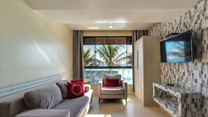 A living room with a sofa, armchair, and TV. A balcony with a view of palm trees is visible through the window. - Pietra Flat (Florianópolis)