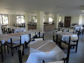 Un comedor amplio con varias mesas y sillas, una barra central, y ventanas grandes que dejan entrar luz natural.