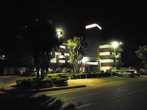 Un edificio de varios pisos iluminado por farolas, rodeado de una zona de estacionamiento.