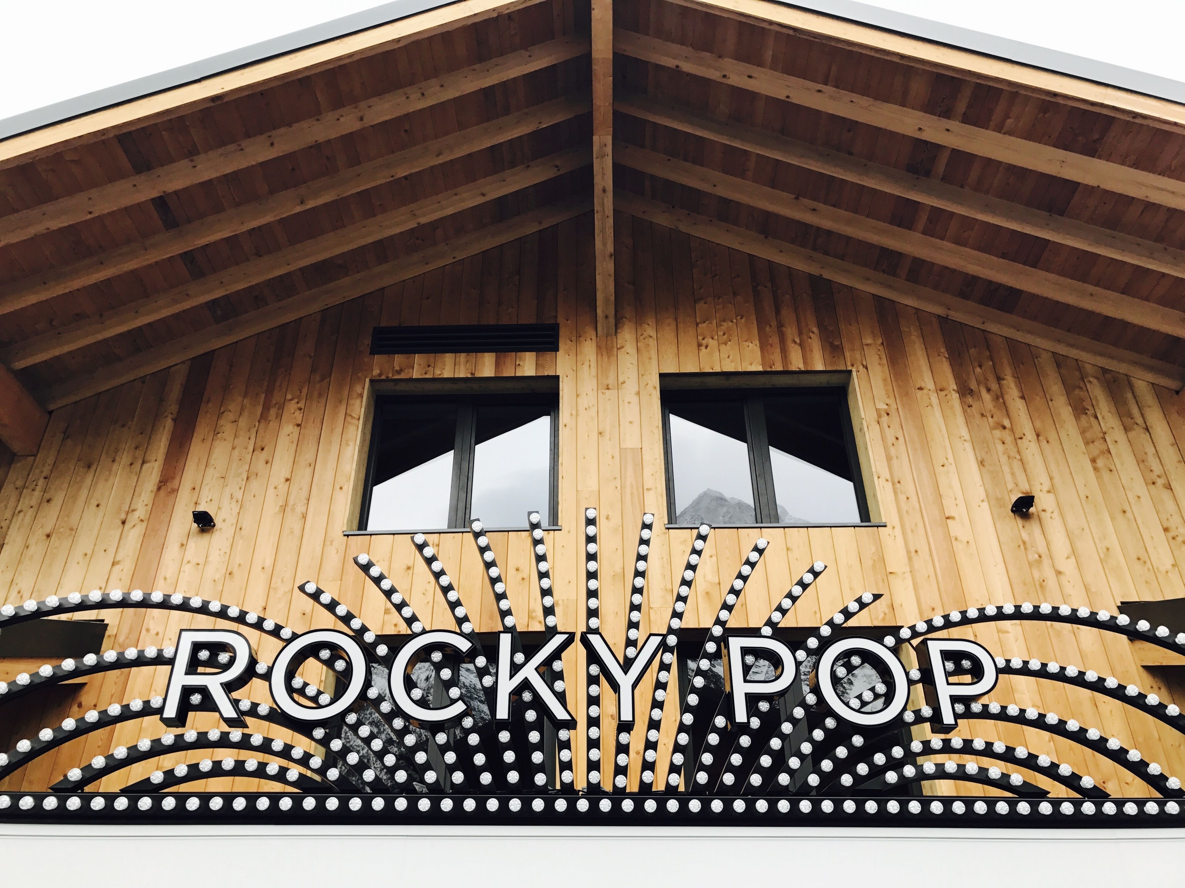 A wooden building with a peaked roof and two windows. The facade has a decorative sign with the word "ROCKYPOP" and a series of lights.