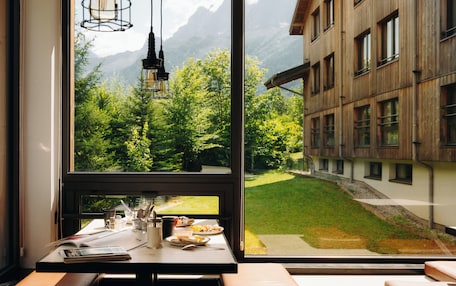 A room with a large window overlooking a mountain landscape, a wooden table with breakfast items, and a view of a building with multiple windows.