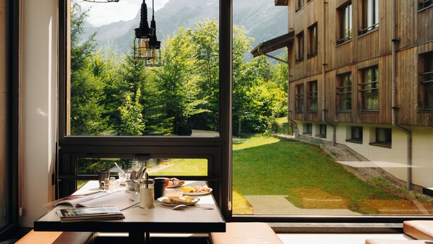 Una habitación con un amplio ventanal que da a un paisaje montañoso, una mesa de madera con provisiones para el desayuno y vistas a un edificio con múltiples ventanas.