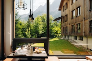 A room with a large window overlooking a mountain landscape, a wooden table with breakfast items, and a view of a building with multiple windows. - RockyPop Chamonix - Les Houches (Les Houches)