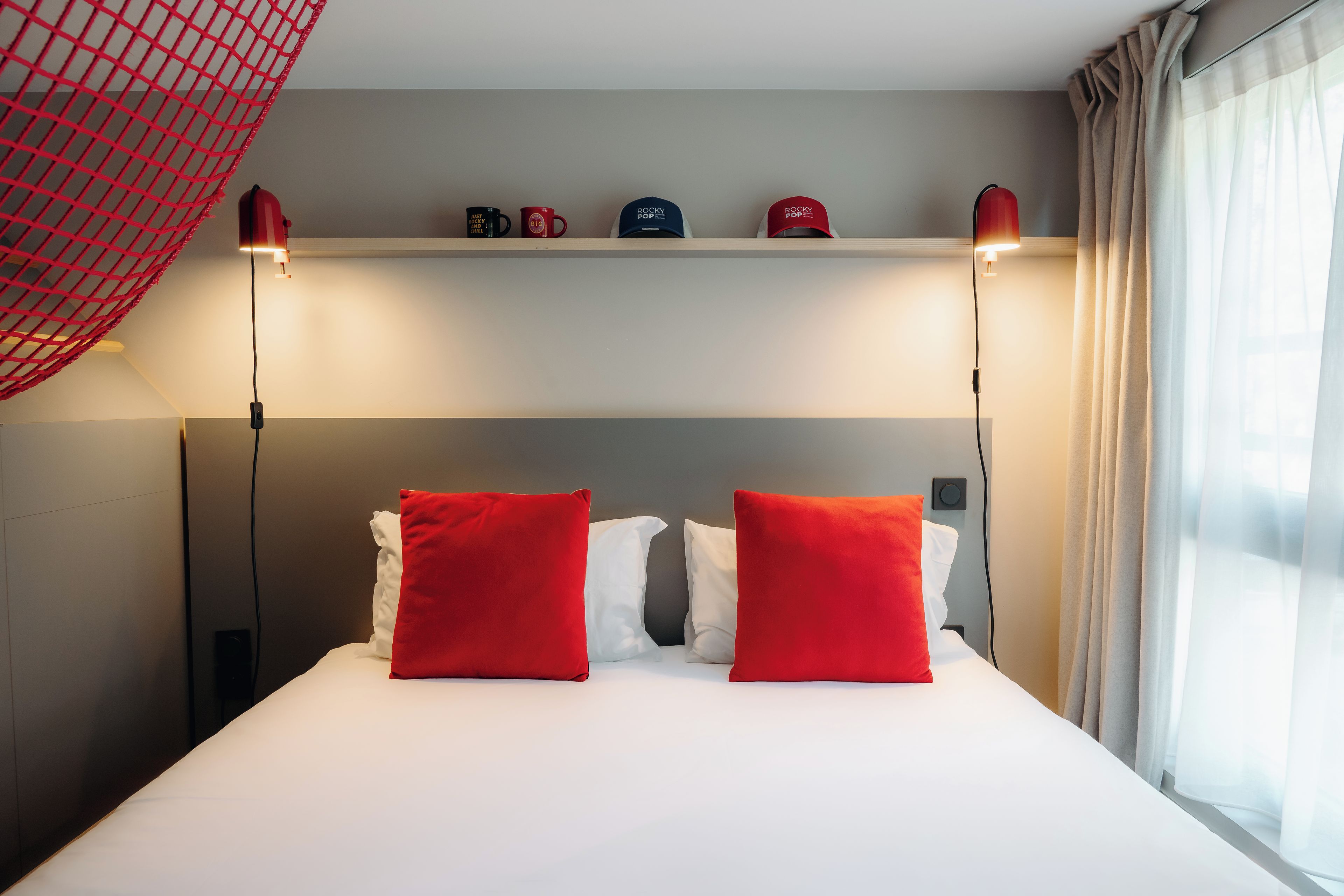 Una camera d'albergo con un letto, due cuscini rossi, uno scaffale con tazze e una finestra con tende.