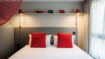 Una camera d'albergo con un letto, due cuscini rossi, uno scaffale con tazze e una finestra con tende.