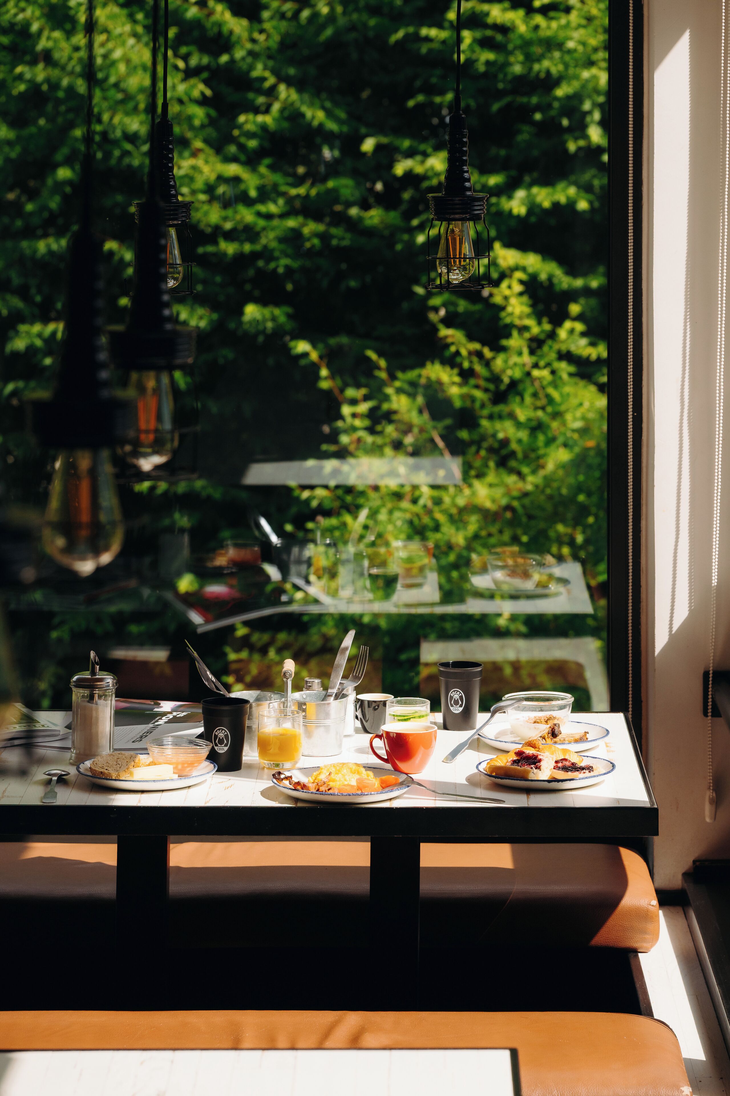 Ein Frühstückstisch mit Tellern, Tassen und einem Saftglas, vor einem Fenster mit Blick auf Grünflächen.