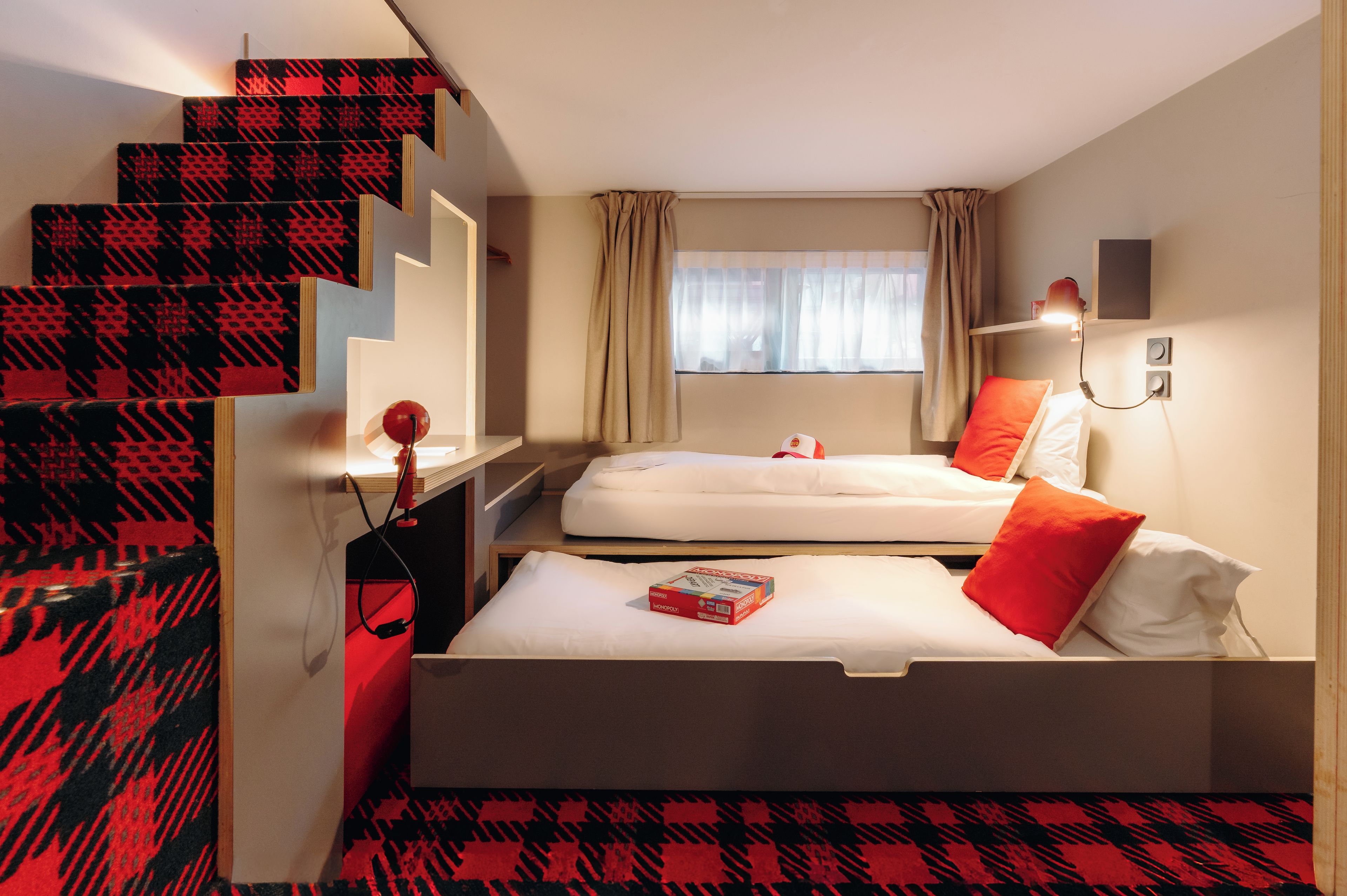 A hotel room with a staircase, two beds, a window with curtains, and a bedside table with a lamp.