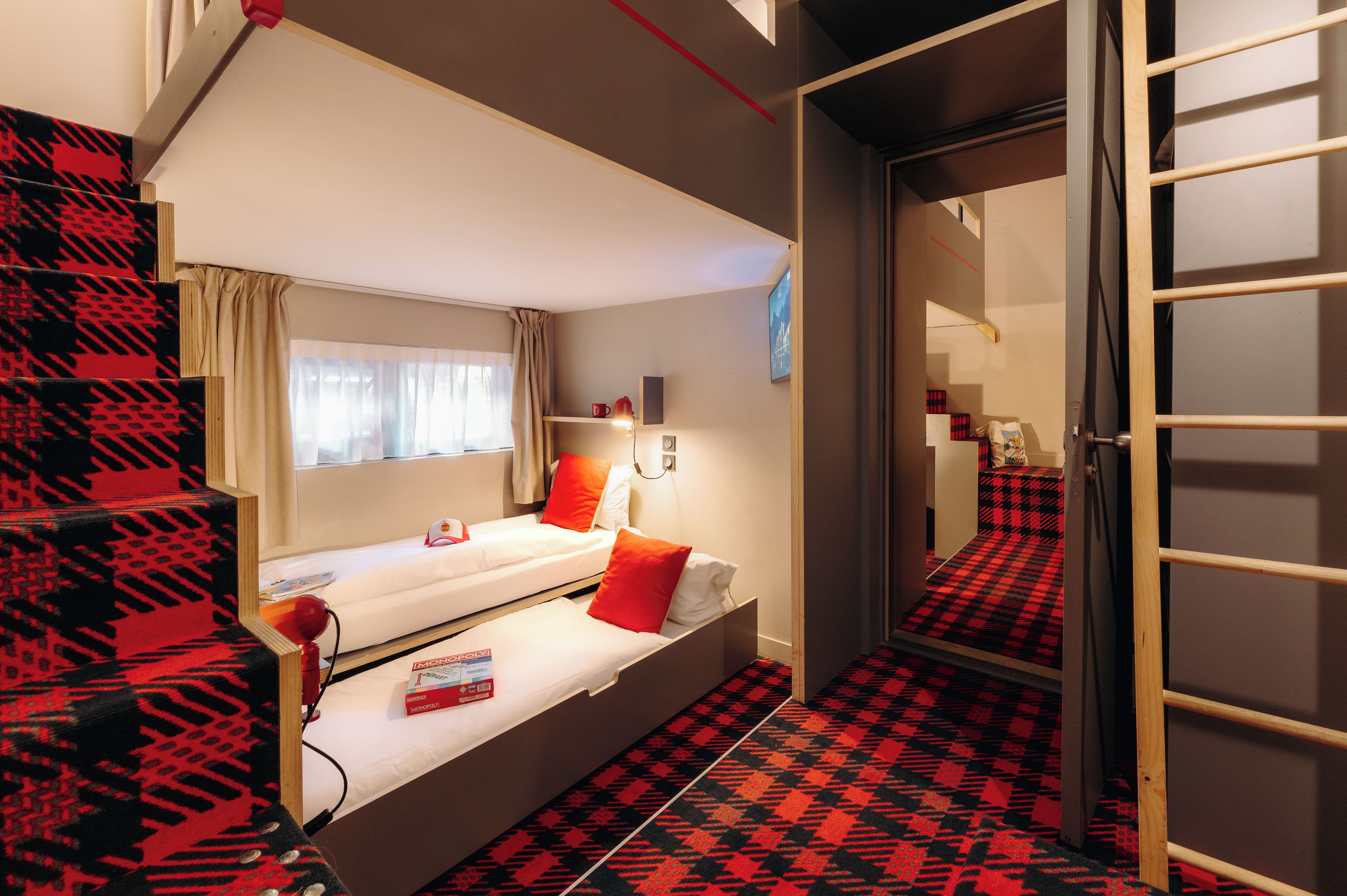 A room with a bunk bed, a small desk, a chair, a window with curtains, and a patterned carpet.