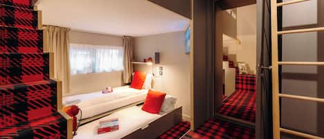A room with a bunk bed, a small desk, a chair, a window with curtains, and a patterned carpet.