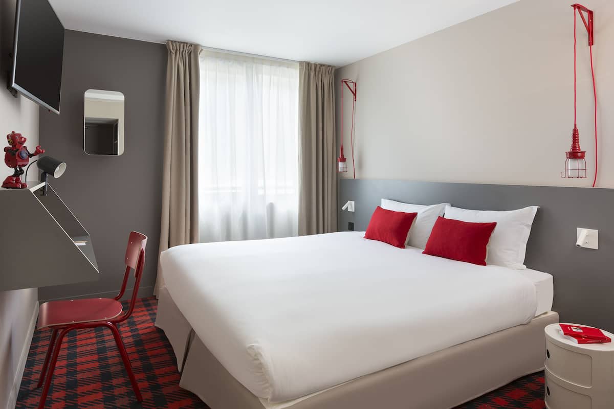 Une chambre d’hôtel avec un grand lit, des oreillers rouges et blancs, une chaise rouge, une petite table et une lampe rouge.