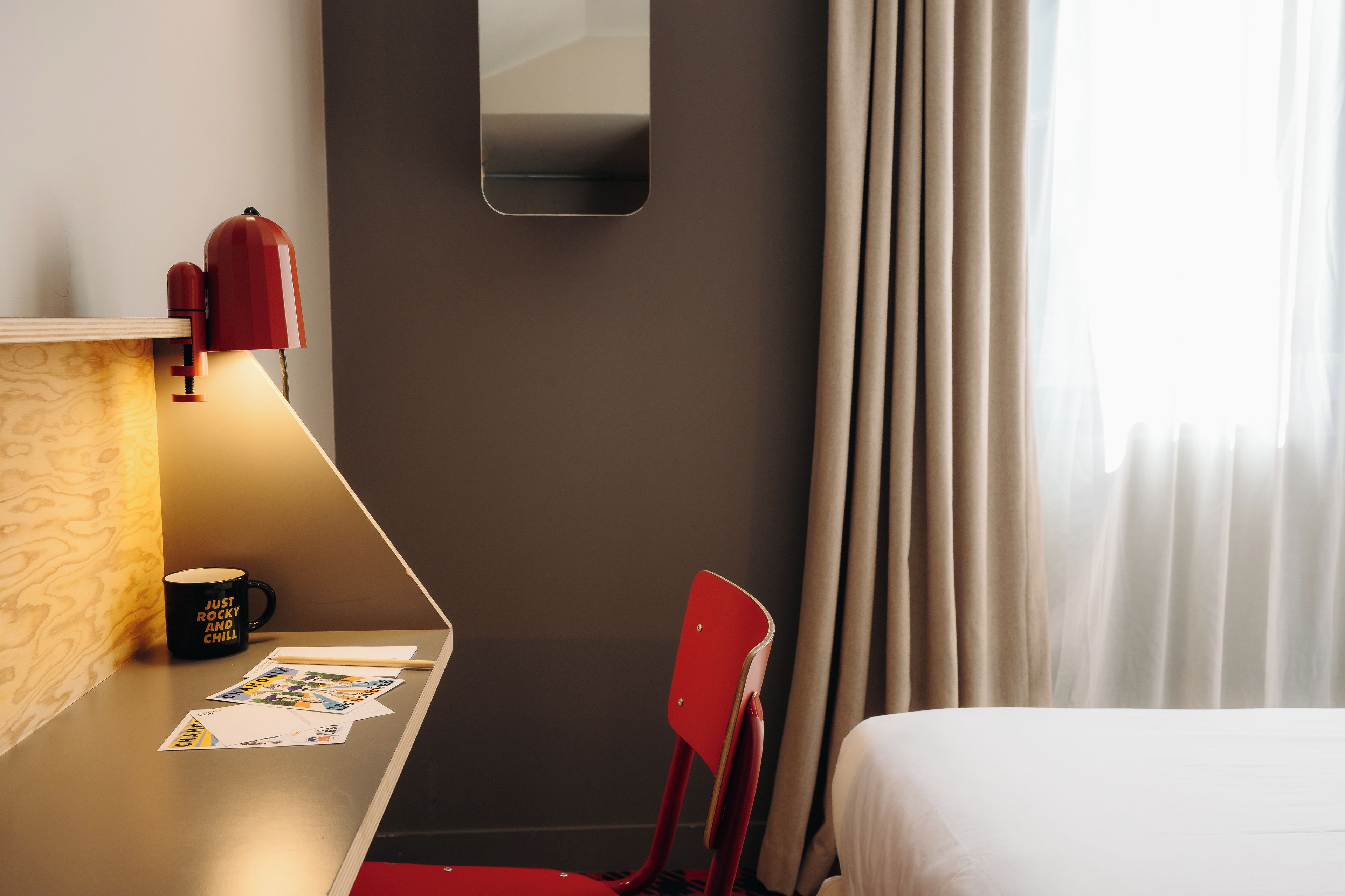A hotel room with a desk, a chair, a bed, a mirror, and a window with curtains.