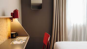 A hotel room with a desk, a chair, a bed, a mirror, and a window with curtains.